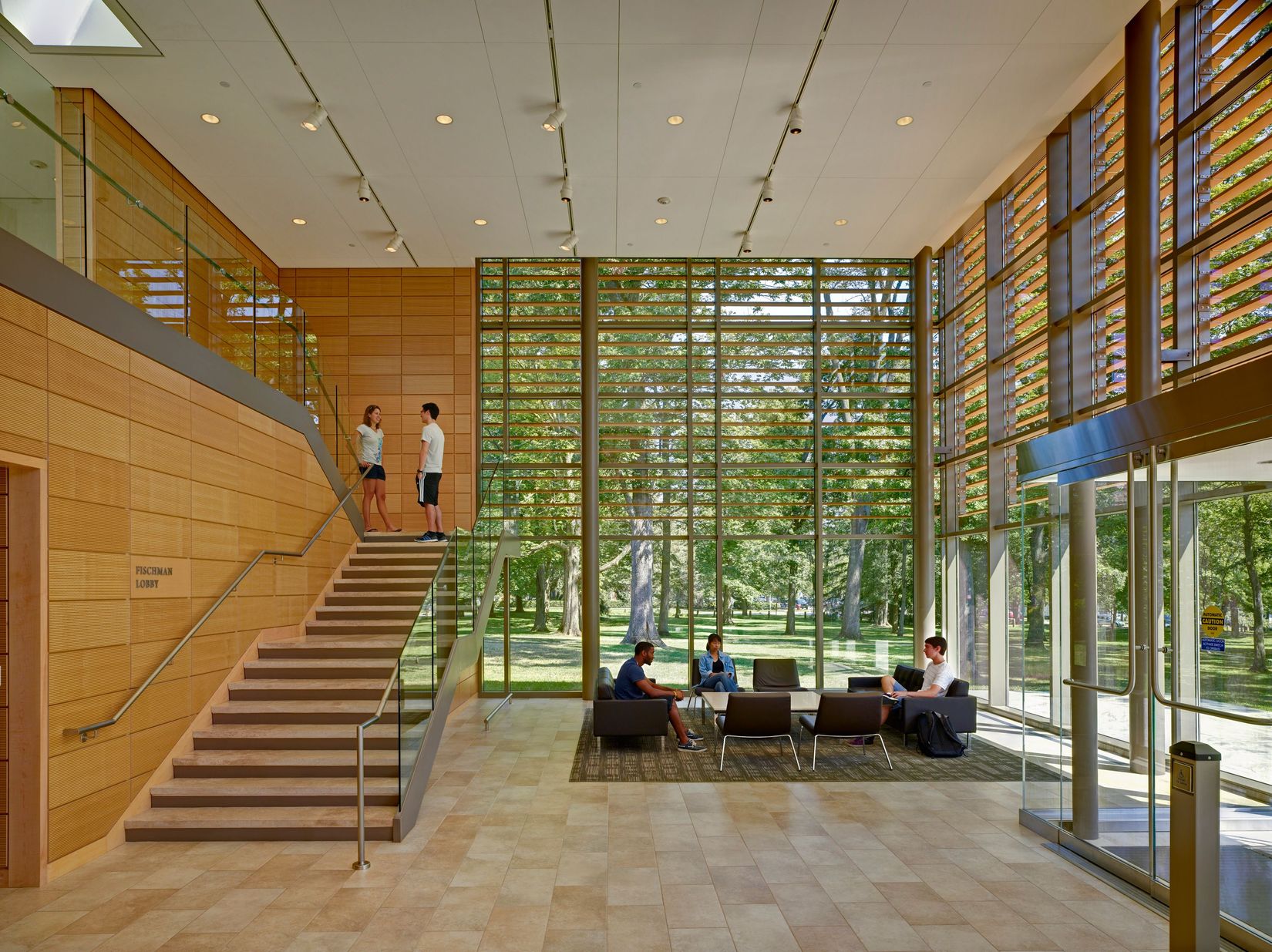 The interior of the Fischman Lobby of Gund Gallery with several people sitting in the seating area and standing on the stair landing. 