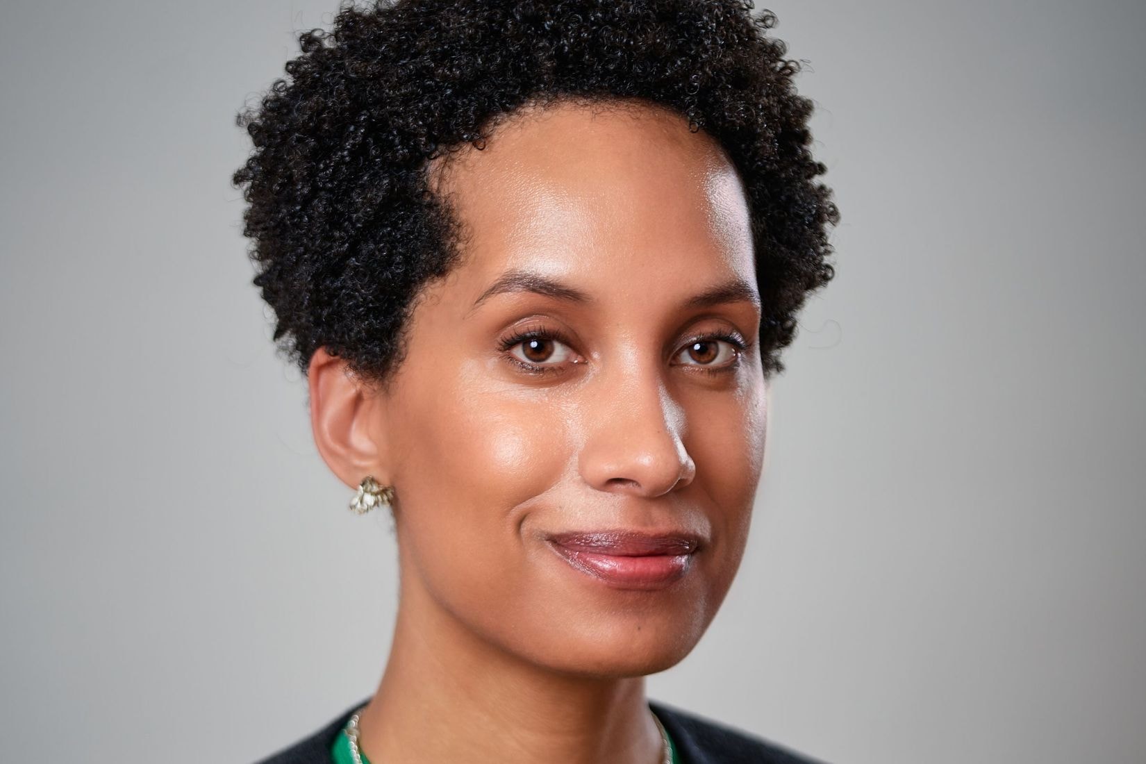A dark complexioned woman with short curly hair smiles wearing a green shirt and dark blazer, and a simple silver necklace.
