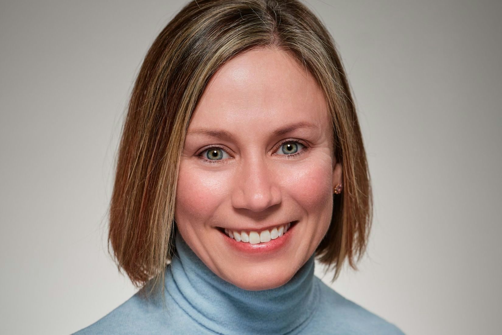 A light skinned woman with chin length dark blonde hair smiles wearing a light blue turtle neck sweater. 