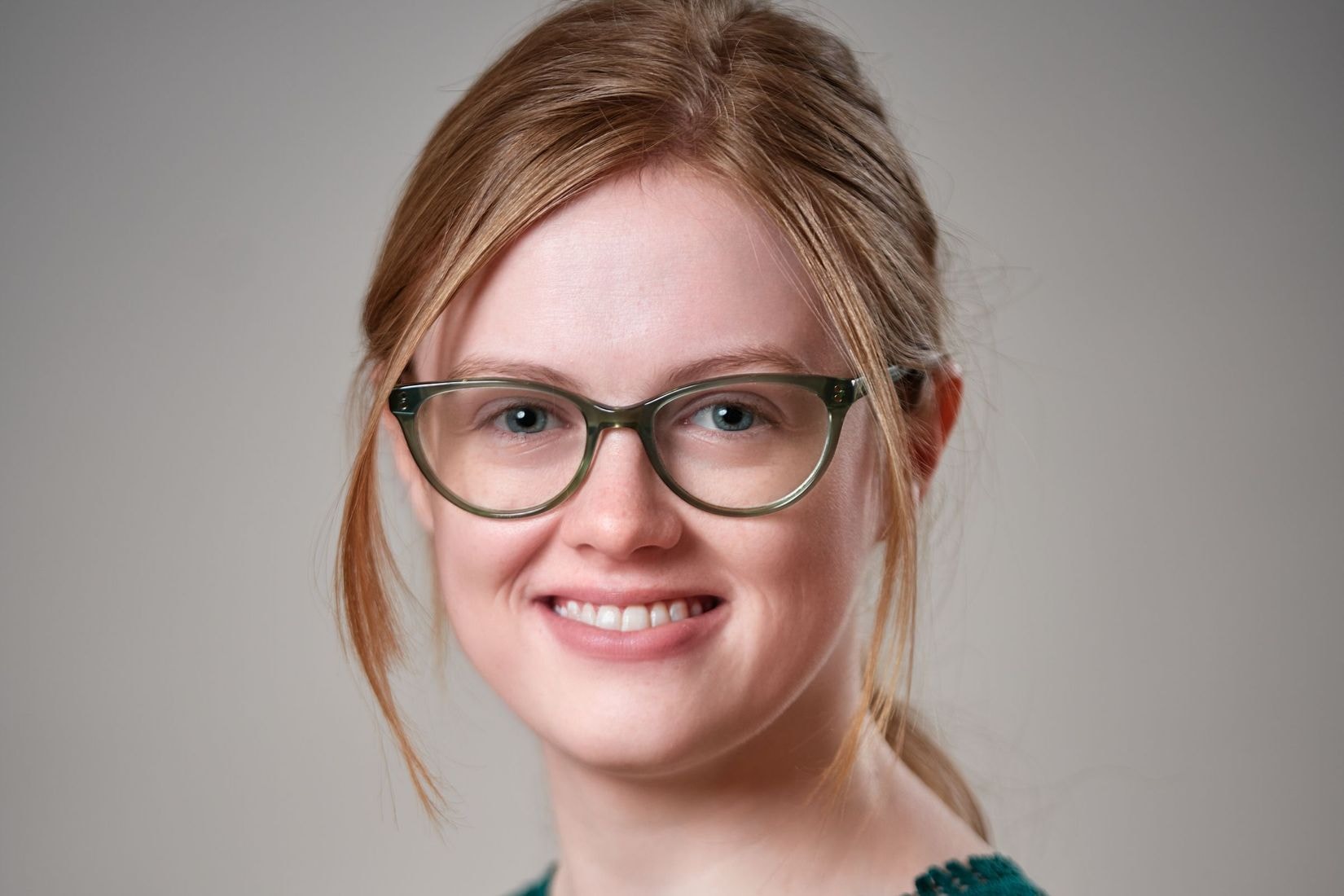 A light skinned woman with strawberry blond hair pulled back in a low ponytail wears a lace emerald green top and glasses.