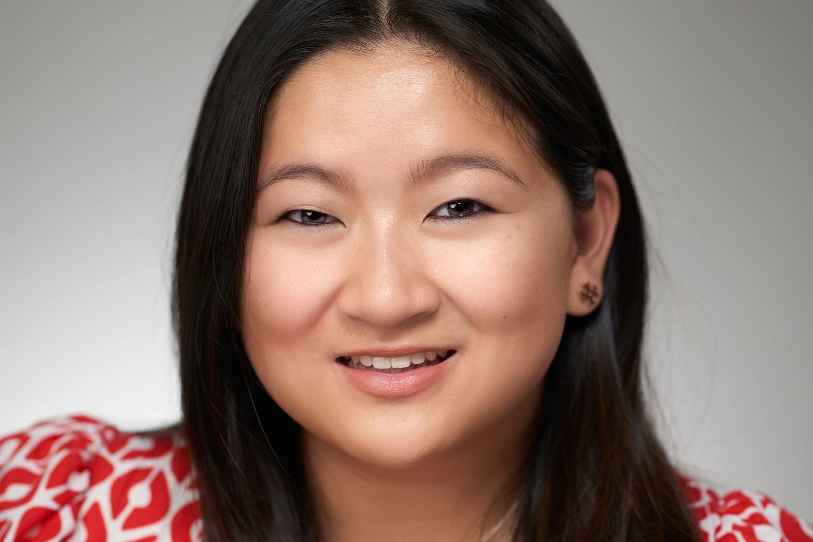 An olive toned woman with dark black hair smiles wearing a red and white patterned shirt, a silver bean necklace, and earrings.