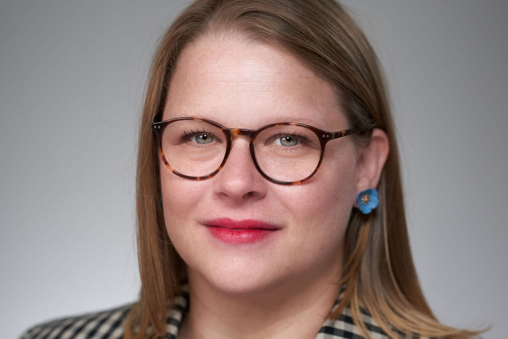 A fair skinned woman with shoulder-length dark blonde hair smiles softly wearing red lipstick, blue flower earrings, a black and white checkered blazer over a black shirt, and tortoise glasses.